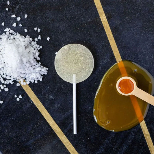 Lollipop with a stick, small bowl of honey, and pile of salt on a dark surface.