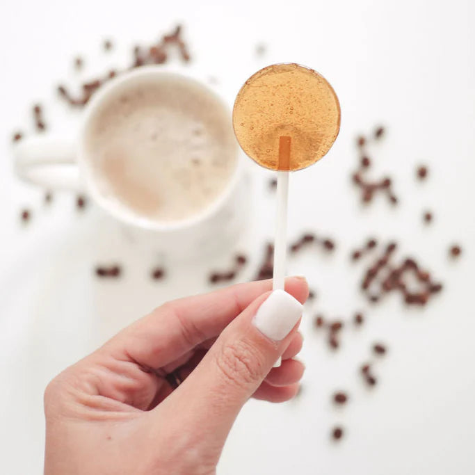 Hand holding a coffee filter on a stick with a blurred cup of coffee and coffee beans in the background.